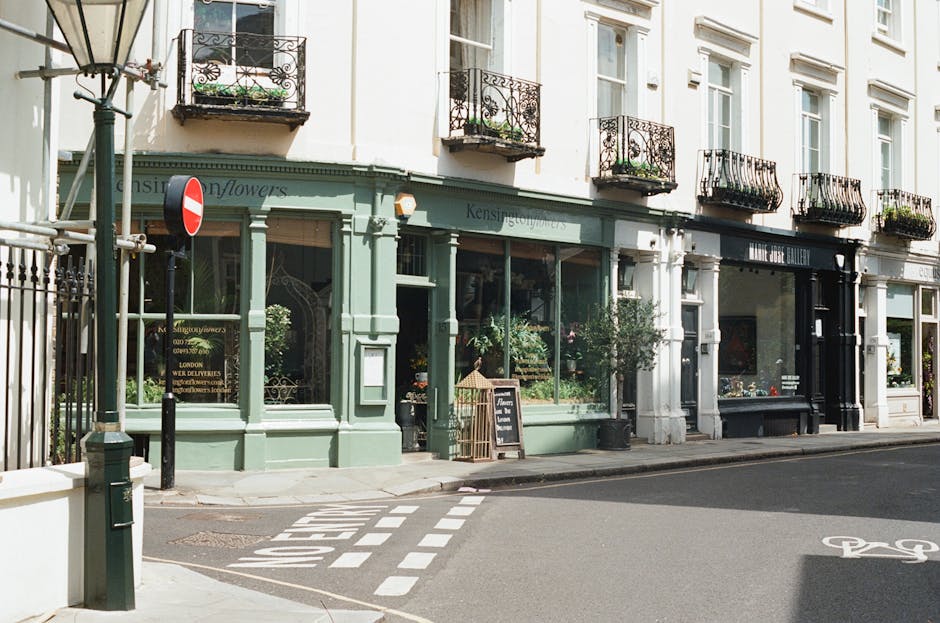 Photograph of a row of retail storefronts on a street in Kensington, featuring large glass display windows, painted in pastel green, black, and white colors. The storefronts include flower shops with visible flower arrangements and decorative plants outside, a chalkboard sign on the sidewalk, and small potted plants near the entrances. The sidewalk is clean and well-maintained, with a bicycle lane marked on the asphalt and a pedestrian crossing. The buildings above have white facades with black wrought iron balcony railings and potted plants on the balconies, illuminated by natural daylight showing a clear, bright day. The scene is calm and tidy, reflecting well-maintained commercial premises in Kensington, aligning with principles of surface cleaning and maintenance, as highlighted in the context of common cleaning mistakes tenants make in Kensington rental flats.