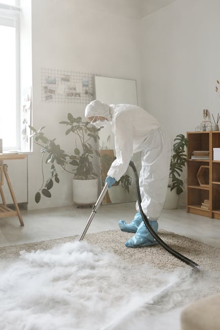 A professional cleaning technician dressed in a white protective suit, gloves, and boot covers is performing deep cleaning on a beige shaggy rug in a bright, modern living room. The technician is using a wet vacuum or extractor machine with a black hose, carefully working on the carpet to remove dirt and moisture. The room features white walls, large windows allowing natural light, and interior decor including potted plants, a wooden shelving unit, and a minimalistic workspace. The surfaces are spotless and well-maintained, demonstrating thorough cleaning and sanitisation. The scene highlights the surface cleaning process essential for domestic cleaning services offered by Carpet Cleaning West Kensington, ensuring hygienic and fresh home environments, particularly for situations requiring emergency flood cleaning or same-day service. The overall atmosphere emphasizes professional and meticulous cleaning practices within a contemporary residential setting.