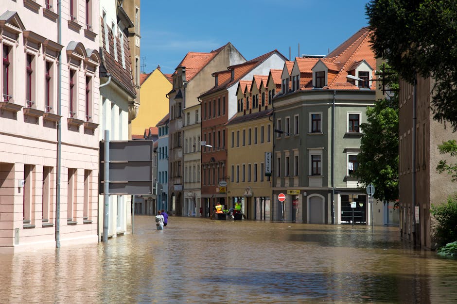 A flooded urban street scene with multi-story residential buildings featuring traditional European architecture, including pastel-colored facades in pink, yellow, green, and beige, and red-tiled sloped roofs. The water level reaches halfway up the first floors, covering the asphalt road and partially submerging parked cars and pedestrians walking through the flood. Bright sunlight illuminates the scene, casting shadows from nearby trees and creating reflections on the water surface. Visible street signs and a traffic light are present, indicating this is a city area. The scene depicts the impact of flooding in Westminster, London, requiring emergency cleaning and flood recovery services, as promoted by Carpet Cleaning West Kensington.
