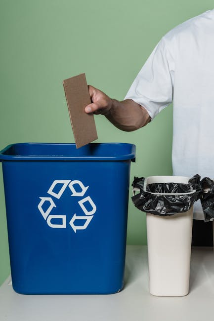 An overflowing urban waste disposal area with multiple large and small bins, including a mixed paper and card bin, a black general waste bin, and a red recycling bin, all surrounded by a considerable 