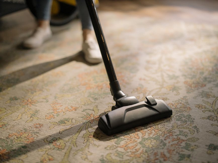 A close-up of a person using a vacuum cleaner with a black head and a long, black hose to deep clean a patterned area rug on a wooden floor in a room with natural lighting. The surface of the rug appears clean and free of dust, with intricate floral motifs in light green, beige, and muted pink tones. The person's lower legs and white shoes are visible in the background, indicating a domestic setting. The scene emphasizes surface cleaning and maintenance, aligning with the upholstery cleaning and stain removal services offered by Carpet Cleaning West Kensington on Kensington High Street.