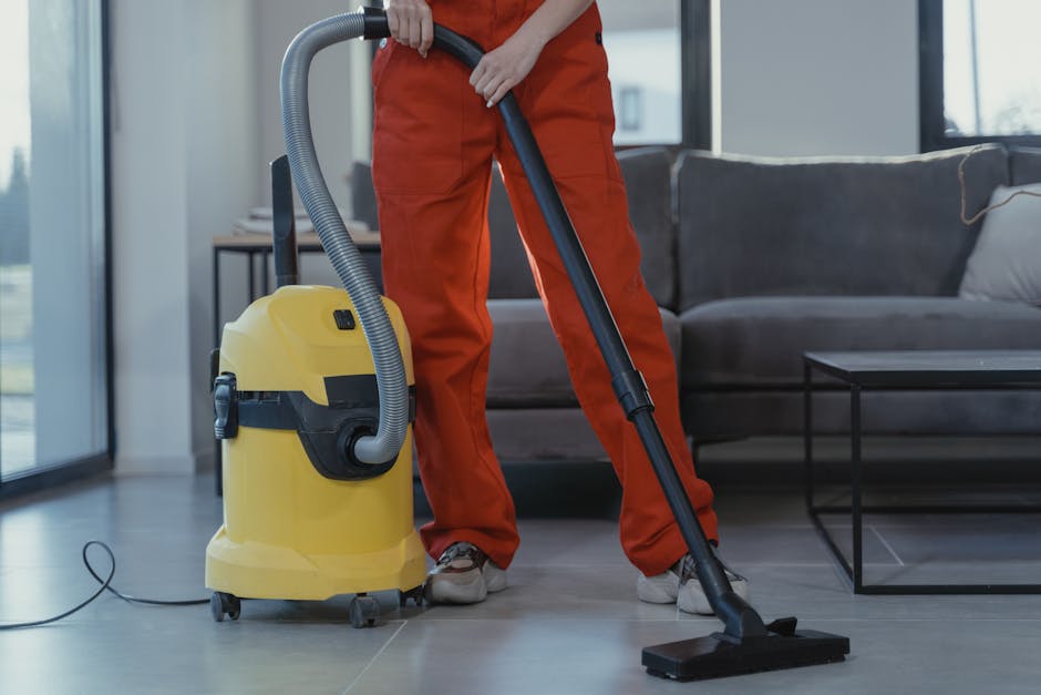 A close-up of a person using a vacuum cleaner with a black head and a long, black hose to deep clean a patterned area rug on a wooden floor in a room with natural lighting. The surface of the rug appears clean and free of dust, with intricate floral motifs in light green, beige, and muted pink tones. The person's lower legs and white shoes are visible in the background, indicating a domestic setting. The scene emphasizes surface cleaning and maintenance, aligning with the upholstery cleaning and stain removal services offered by Carpet Cleaning West Kensington on Kensington High Street.