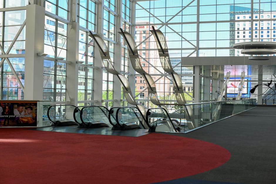 Inside a modern exhibition venue with large floor-to-ceiling glass windows revealing a cityscape of tall buildings beyond. The interior features escalators and a wide open space with a red carpeted area and dark tiled flooring. The escalators are clean and shiny, positioned side by side, leading to a mezzanine level. Natural light floods the space, emphasizing the bright and pristine condition of the surfaces. The environment appears well-maintained, aligning with professional deep cleaning and surface sanitisation services promoted by Carpet Cleaning West Kensington.