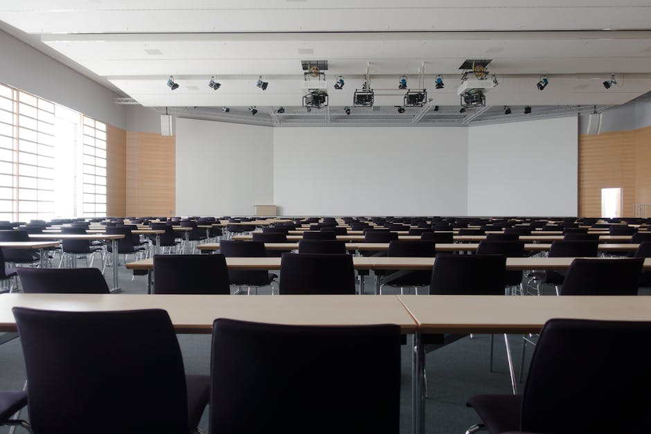 A spacious conference room with a high ceiling and large windows on the left allowing natural light to brighten the space. The room is furnished with multiple rows of rectangular tables, each with a smooth, light-colored surface, and black upholstered chairs arranged neatly around them. The floor appears to be covered with dark carpeting, and the walls are painted in neutral tones, with some wooden panel accents on the sides. The ceiling is equipped with several mounted professional lighting and audiovisual equipment, including ceiling speakers and cameras, indicating that the room is used for presentations or events. The area looks clean, well-maintained, and ready for an event, reflecting professional surface cleaning and maintenance services provided by Carpet Cleaning West Kensington, as featured on the page about Olympia exhibition venue carpet cleaning for event organisers.