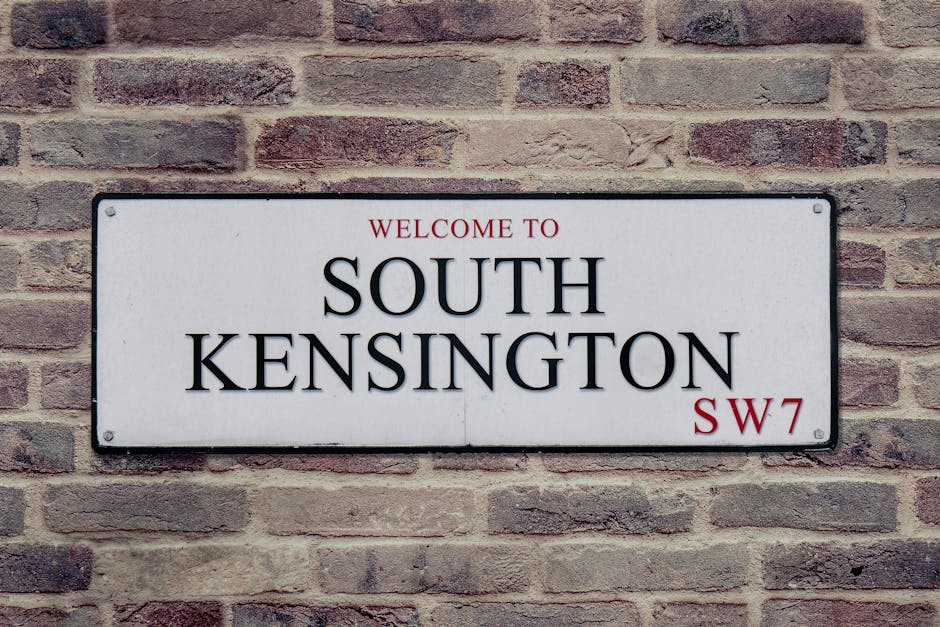 A metal street signpost with three rectangular signs mounted vertically, situated outdoors among lush green leafy trees. The top sign is red and white, indicating tow-away and no stopping regulations 
