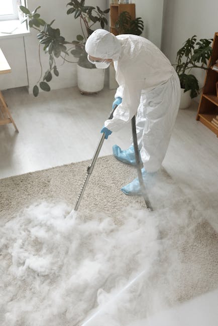 A professional cleaner wearing a full white protective suit, gloves, and a face mask is using a steam cleaning machine to deep clean a beige carpet in a modern, well-lit living room. The room features