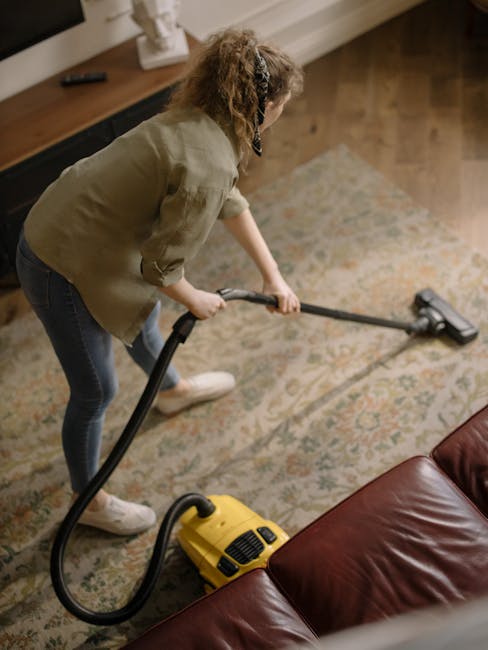 A yellow vacuum cleaner with a flexible black hose and a metal extension tube is positioned on an ornate floral-patterned rug in a room with wooden flooring. The vacuum cleaner is plugged into an outlet and appears to be used for surface cleaning or deep cleaning of the carpet. Behind the rug, there is a black metal and wooden console table with a remote control, stacked papers, and a decorative bust on top. The room has white skirting boards and a corner of a piece of wooden furniture visible in the background, with natural lighting enhancing the clean and organized appearance, typical of residential or commercial cleaning environments, related to surface sanitation and maintenance in West Kensington flats.